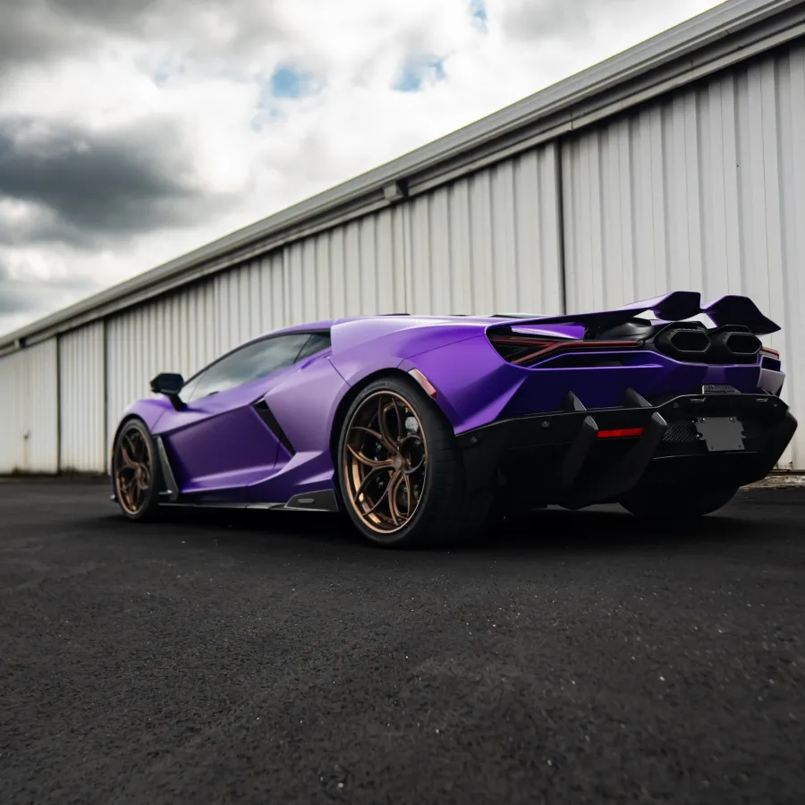 Purple sports car with bronze wheels parked on black asphalt near industrial building under cloudy sky.