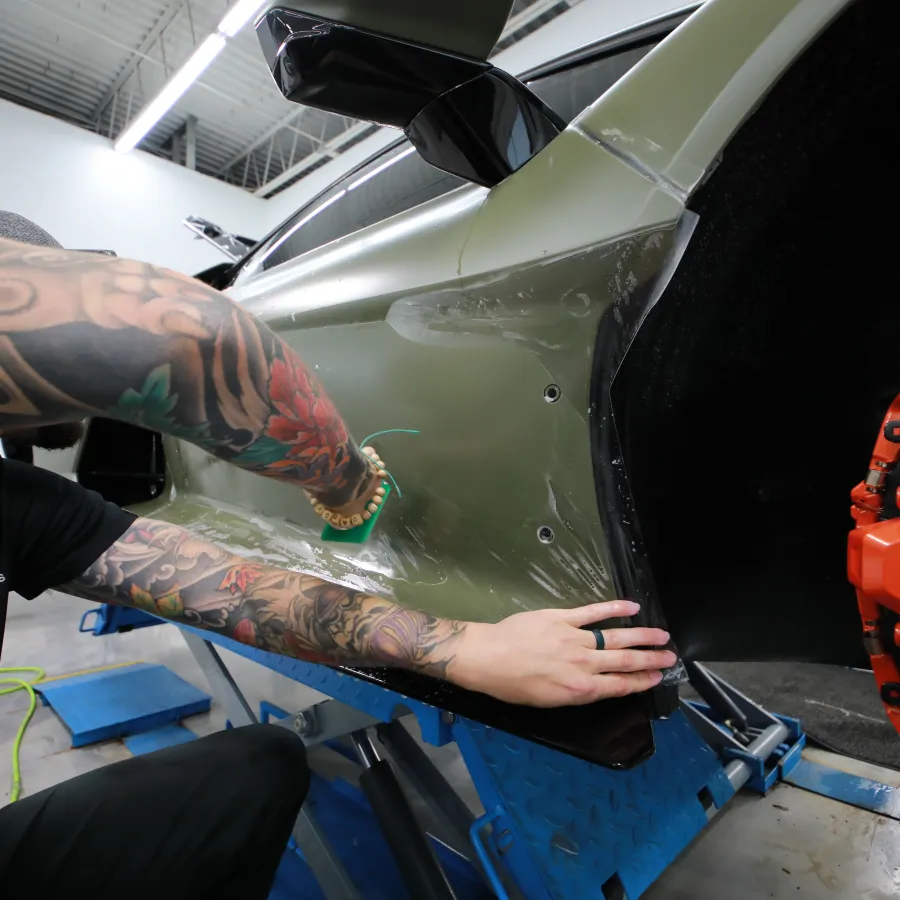 Tattooed technician installing protective film on matte olive Lamborghini car in workshop with orange brake caliper visible.