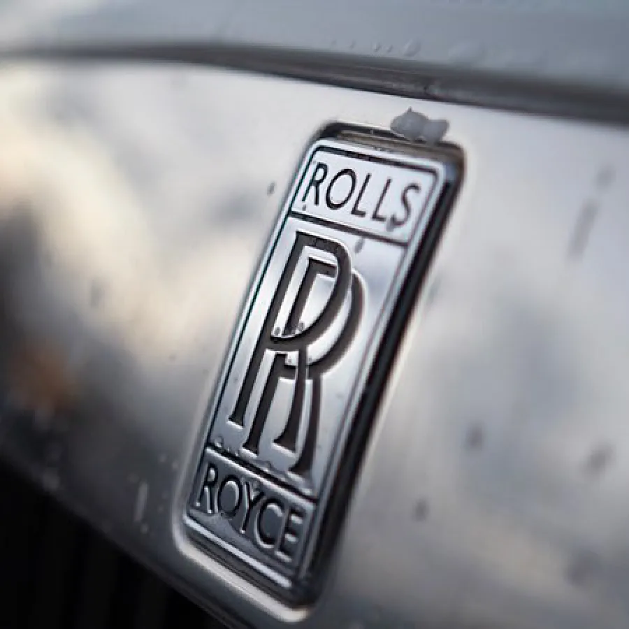 Close-up of a Rolls-Royce logo on a shiny silver car grille with reflections and blurred background.