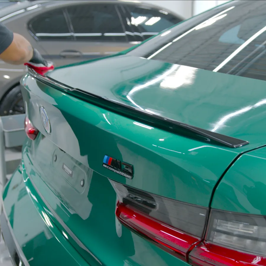 Person applying paint protection film on the rear trunk of a green BMW M5 in a workshop.