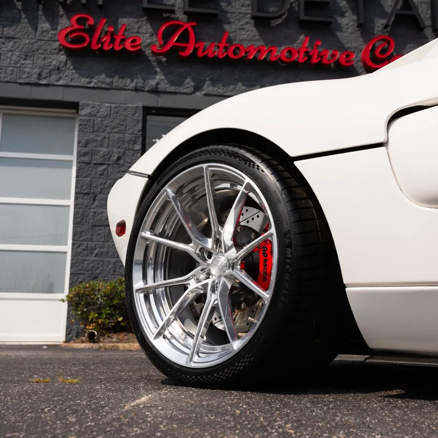Close-up of a white sports car's polished alloy wheel and red brake caliper outside Elite Automotive shop.