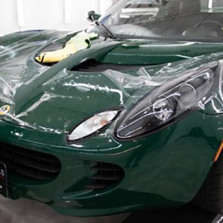 Person applying clear protective film on the hood of a dark green sports car in a workshop.