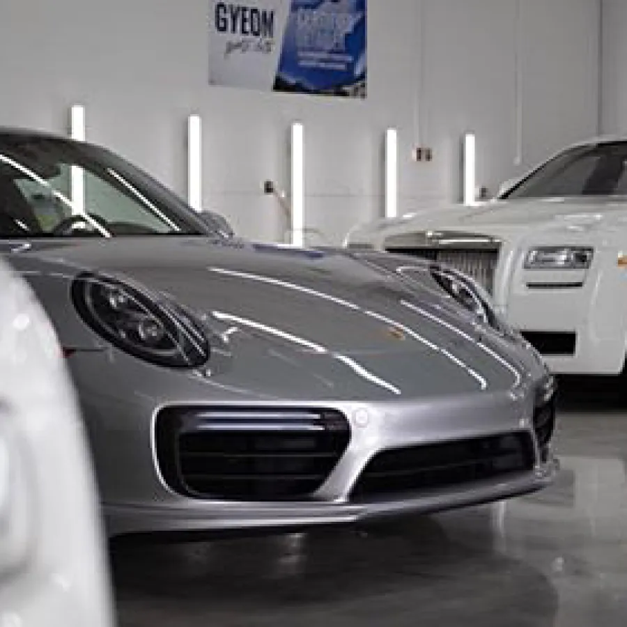 Silver Porsche and white Rolls-Royce luxury cars parked indoors on shiny floor with bright lighting.