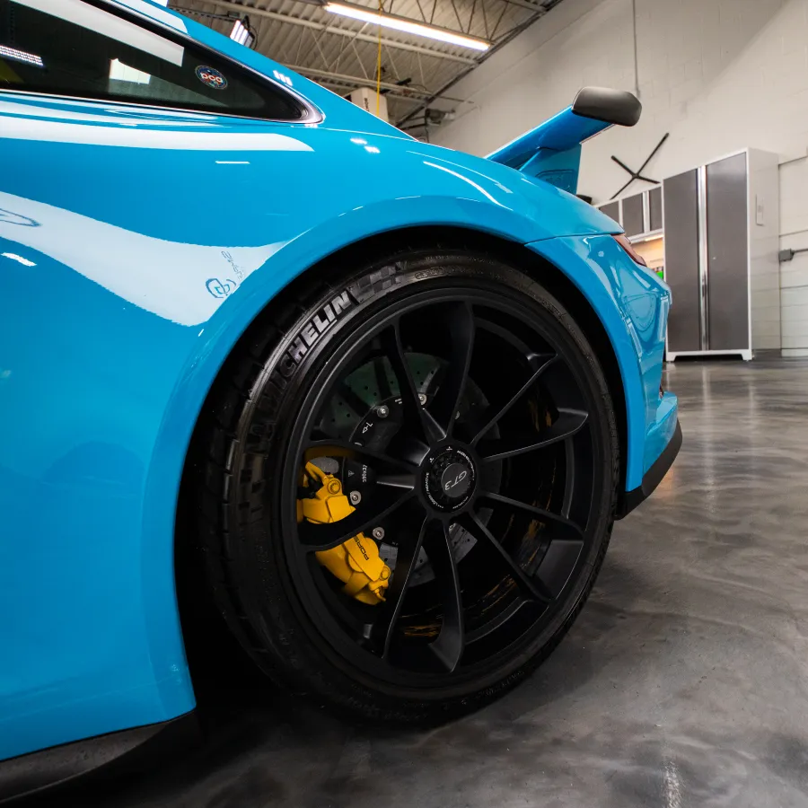 Close-up of a blue sports car’s rear wheel with black rims and yellow brake calipers in a garage.