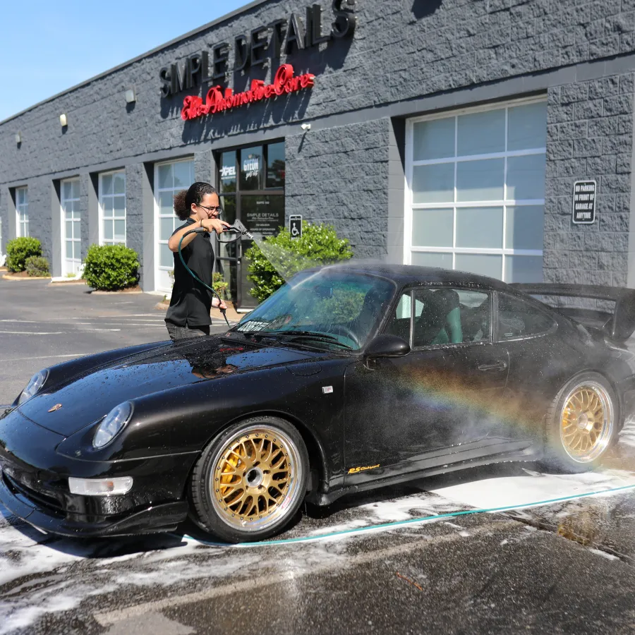 Person washing a black Porsche with gold rims outside Simple Details auto care under bright sunlight.
