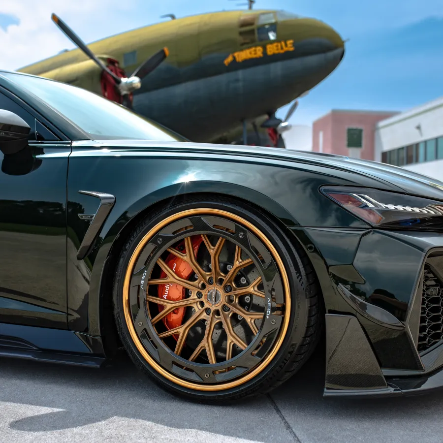 Close-up of a luxury black sports car with gold-accented rims parked near a vintage airplane and modern buildings.