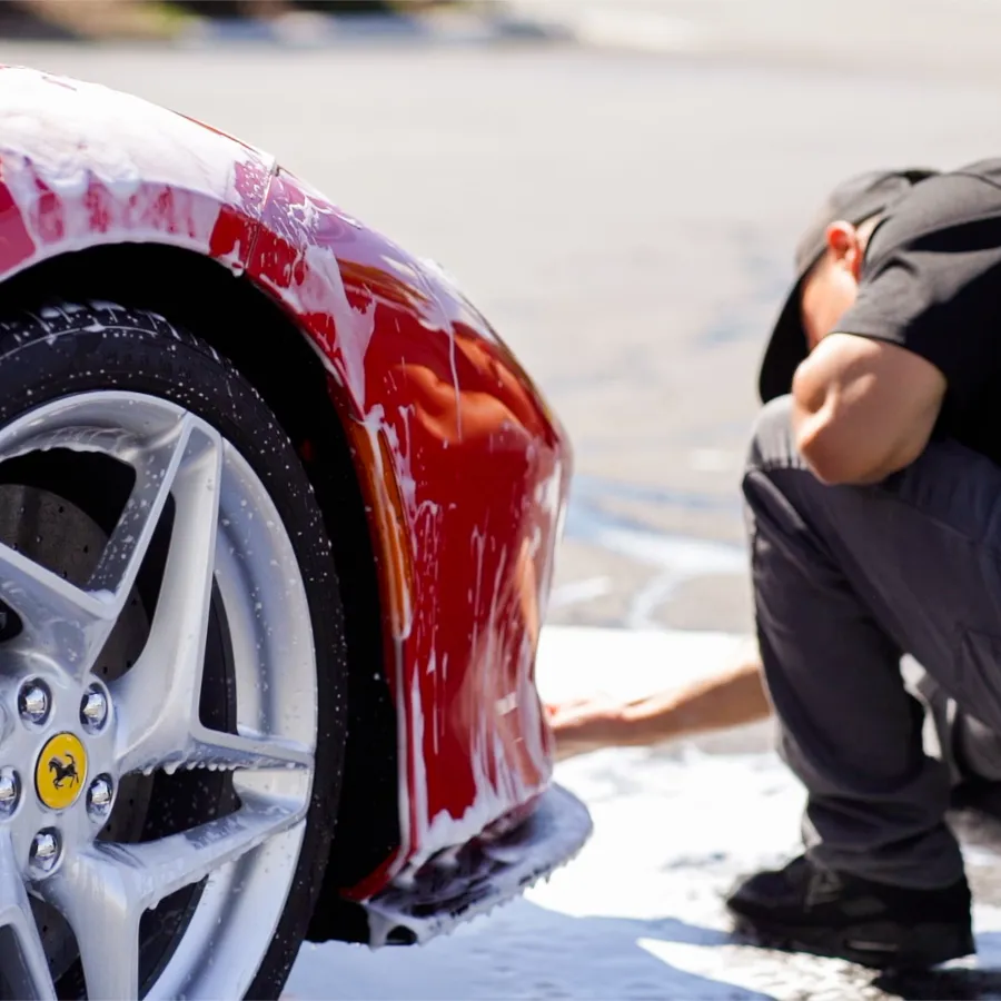 Person washing a red Ferrari sports car with soap foam on a sunny day outdoors.