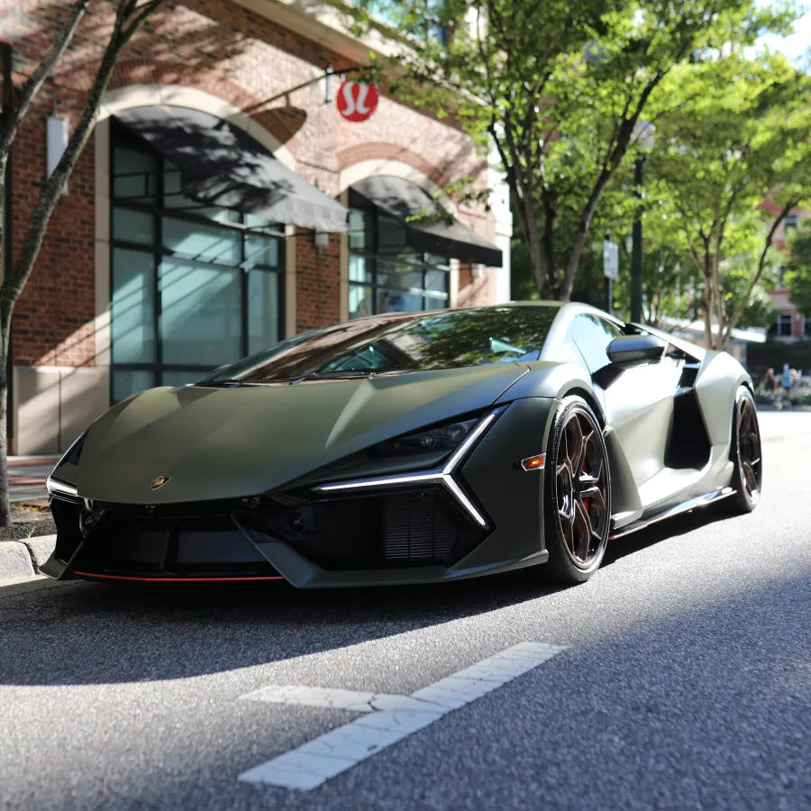 Matte green Lamborghini sports car parked on a city street near brick storefront with trees and sunlight.