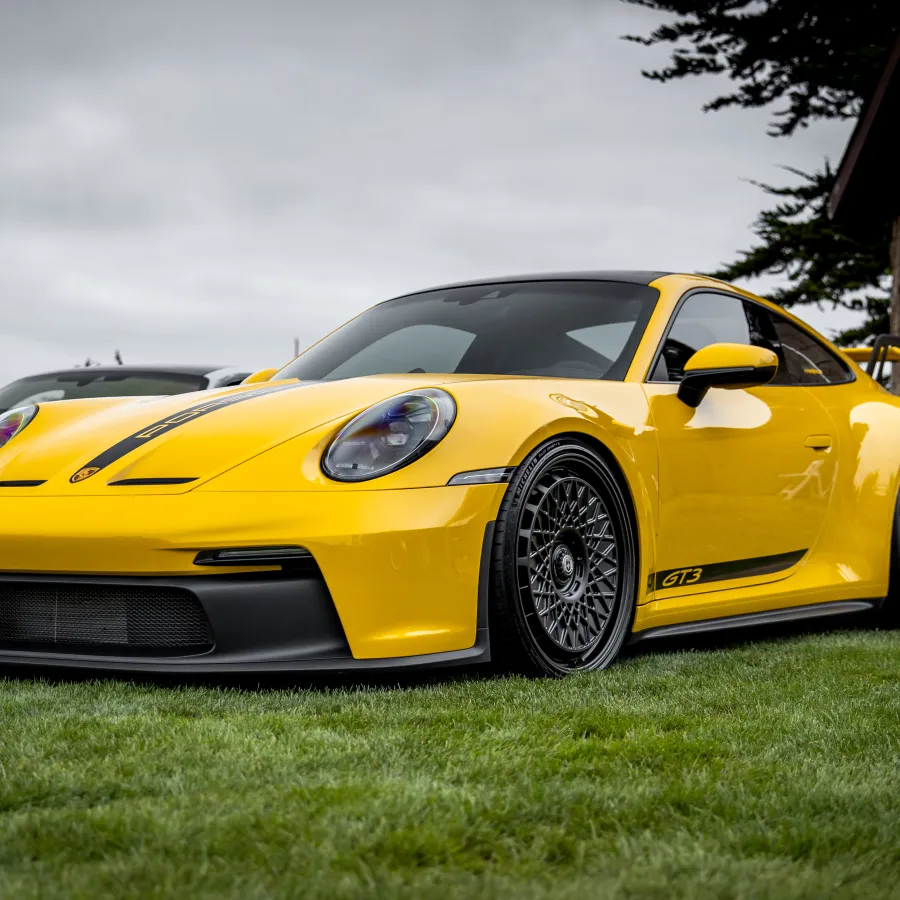 Yellow Porsche GT3 sports car with black trim and mesh wheels parked on grass under cloudy sky.