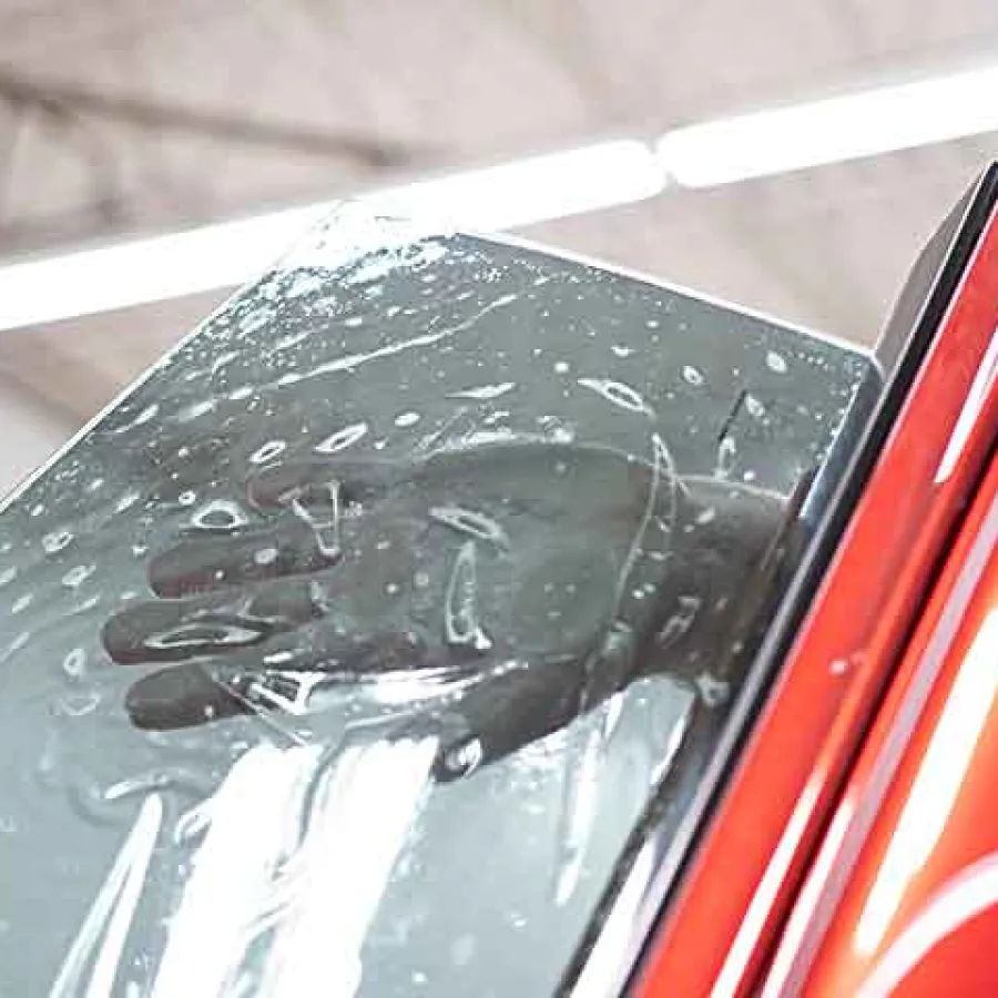 Hand applying clear protective film onto red car door window in a bright garage setting
