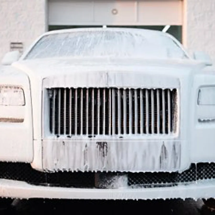 White luxury car covered in foam during a wash in front of a garage building.