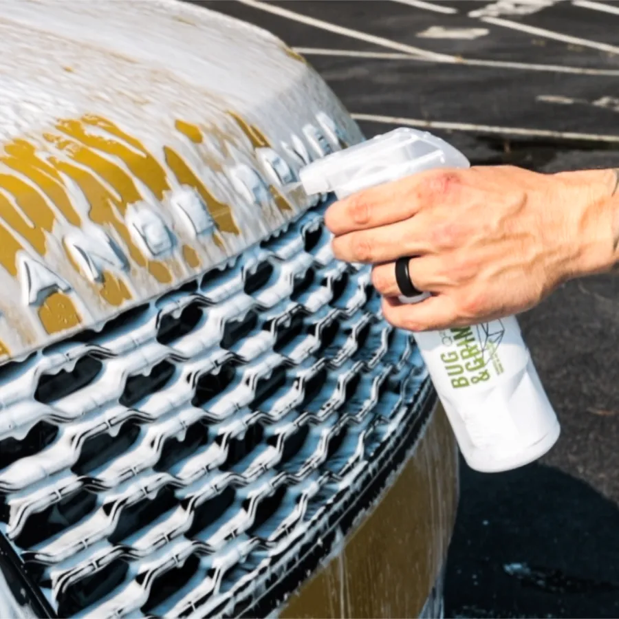 Person spraying foam cleaner on a golden car grille for washing and detailing outdoors in a parking lot.
