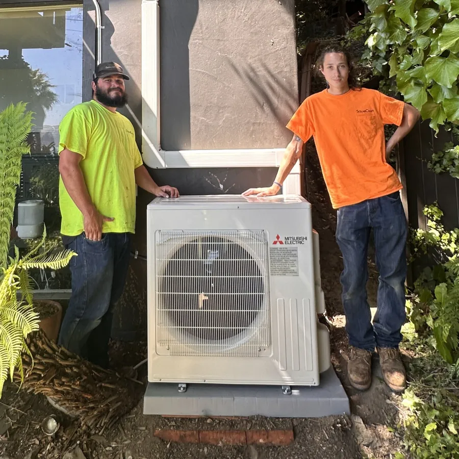 Two workers standing beside a newly installed Mitsubishi Electric outdoor HVAC unit outside a house surrounded by plants.