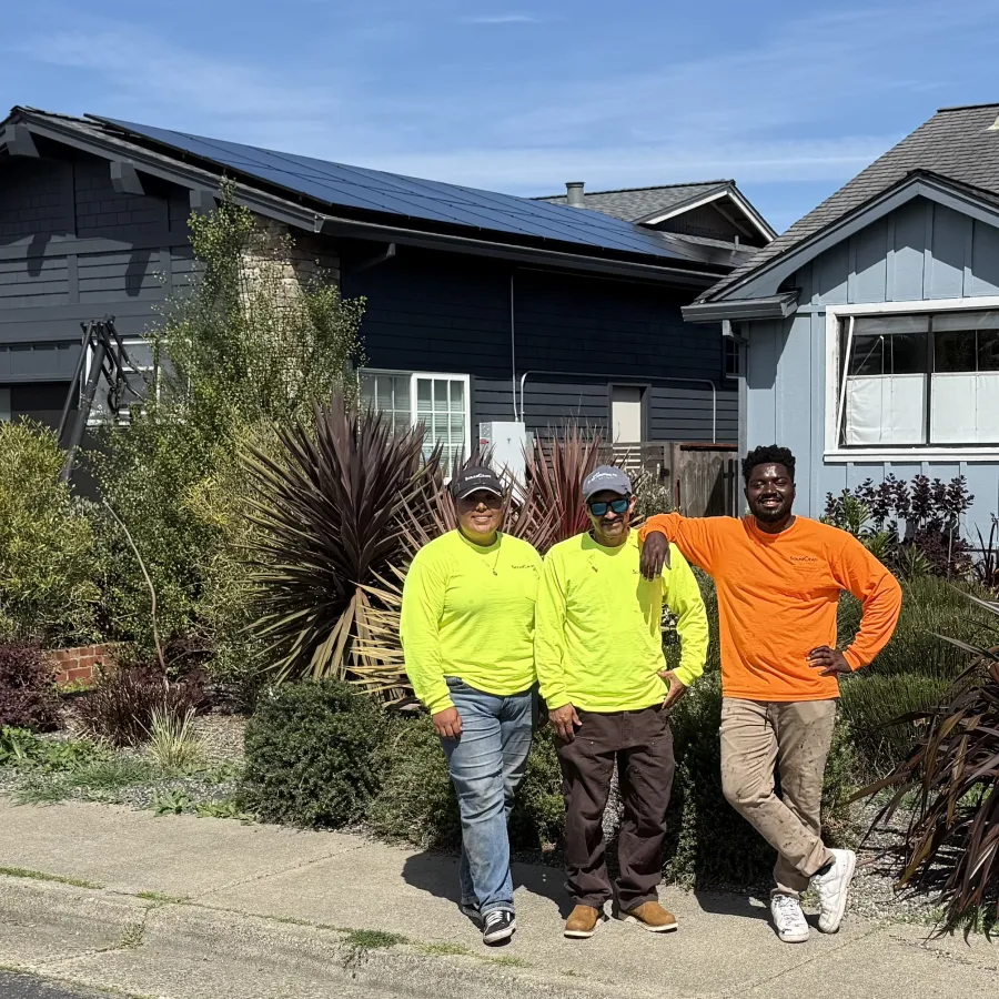 Three workers in bright safety shirts standing on a sidewalk by houses with garden plants and solar panels on the roof in larkspur