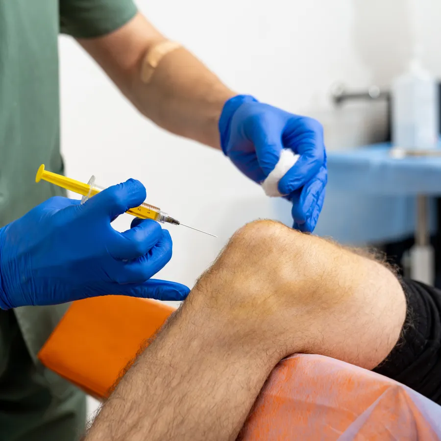 Medical professional with blue gloves administering a knee injection to a patient resting on an orange chair.