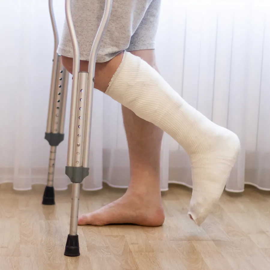 Person with a cast on foot using crutches standing on wooden floor in front of sheer curtains