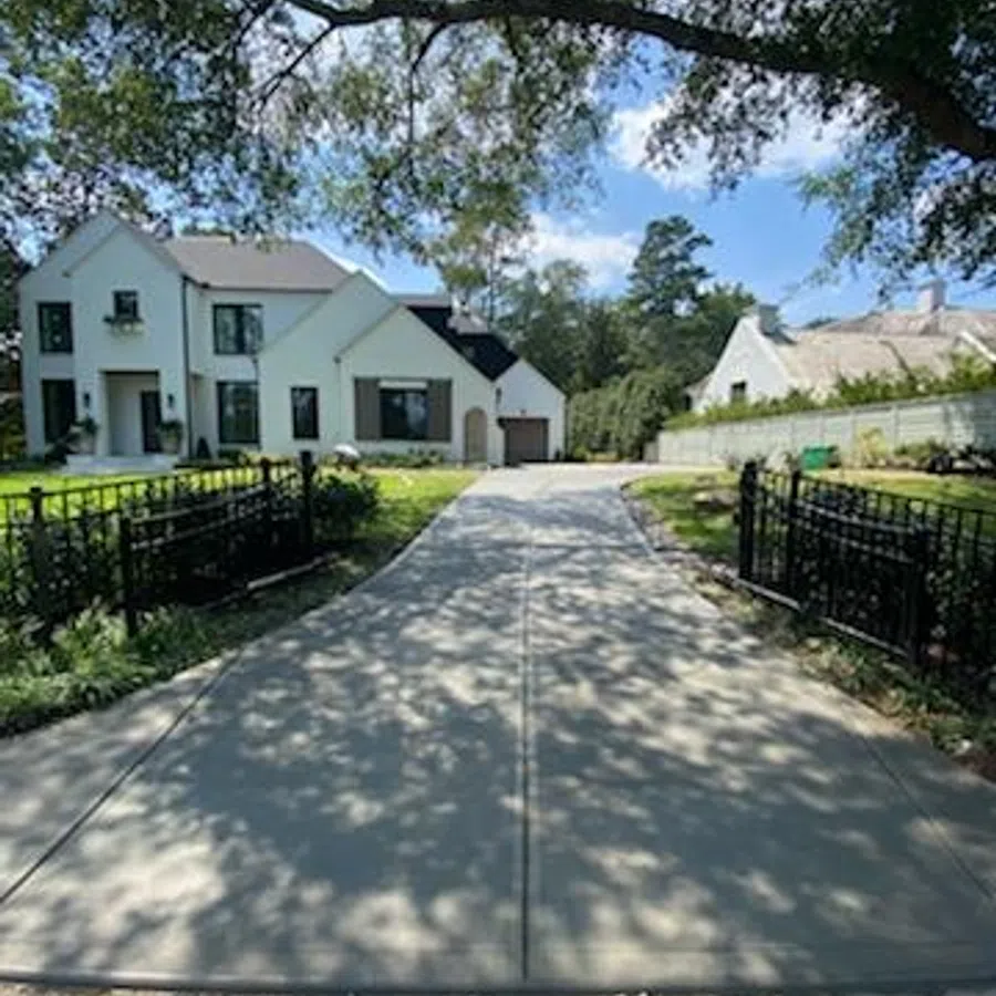 Long driveway leading to a modern white house with black trim, surrounded by green trees and a sunny sky.