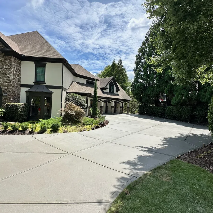 Spacious driveway leading to a large house with stone and stucco exterior surrounded by greenery under a partly cloudy sky