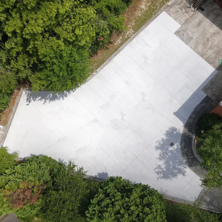 Aerial view of a large, empty white concrete driveway surrounded by green trees and shrubs.