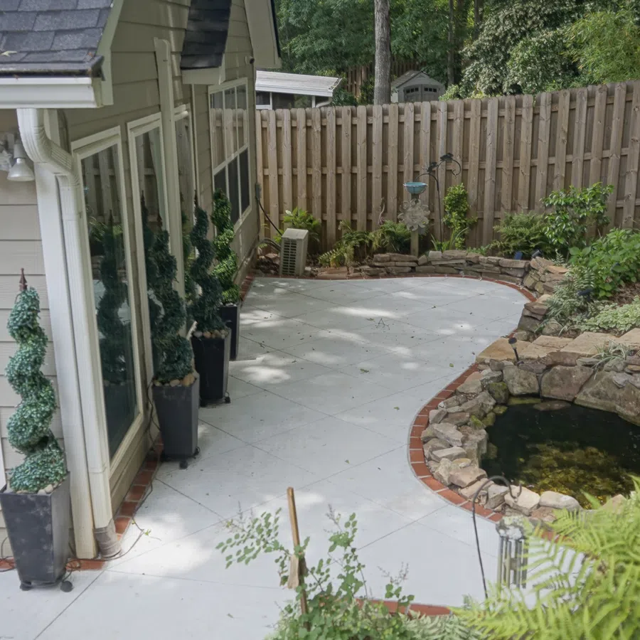 Curved backyard patio with potted plants, stone pond, wooden fence, and lush greenery surrounding the area.