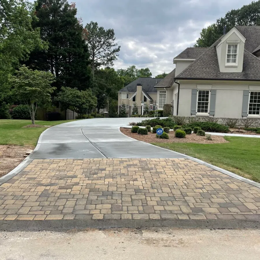 Curved concrete and stone brick driveway leading to a white suburban house with manicured lawn and landscaping.