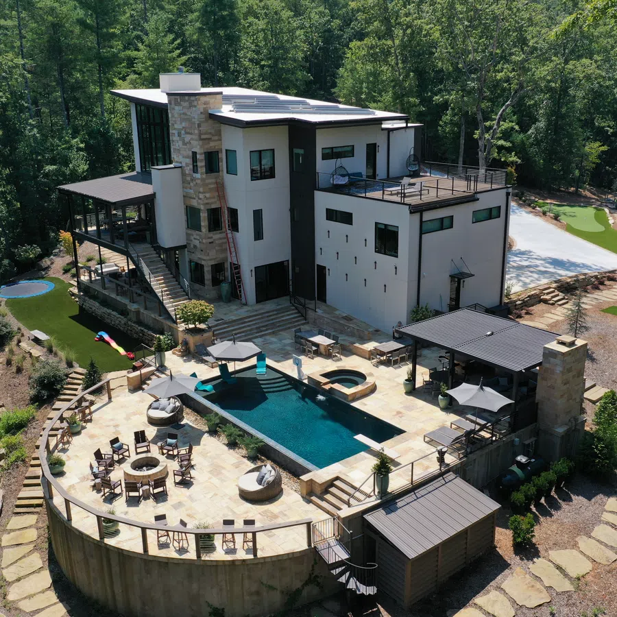 Modern luxury home surrounded by forest featuring a pool, outdoor seating, fire pits, and tennis court under clear sky.