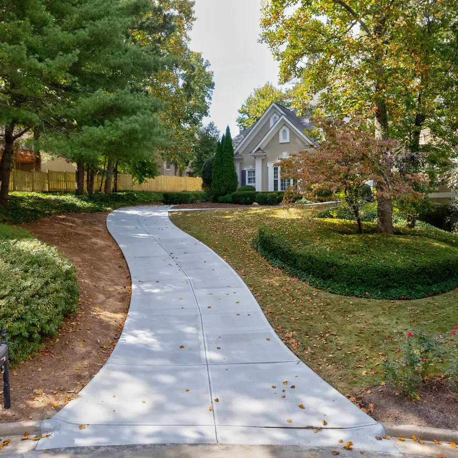 Curved concrete driveway leading to a charming house surrounded by trees and well-maintained landscaping.