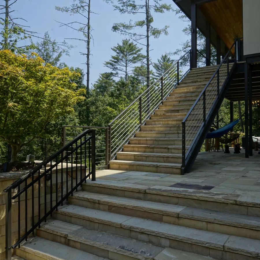 Outdoor natural stone staircase with black metal railings leading to covered outdoor living space surrounded by trees.