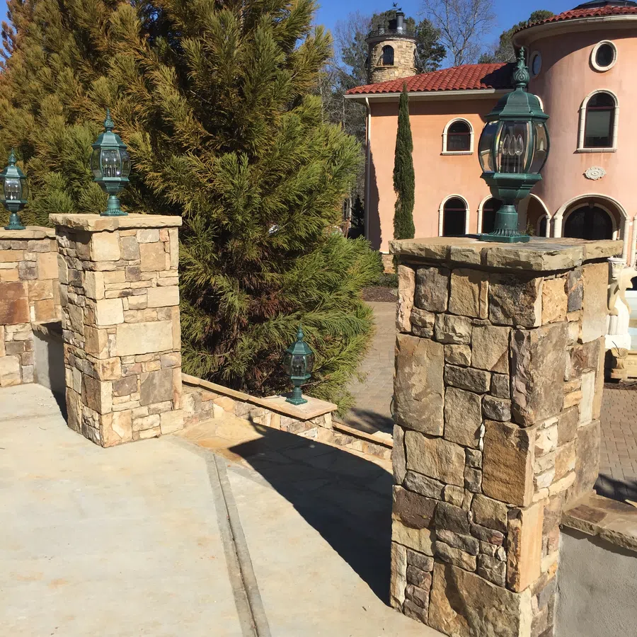 Stone pillars with vintage lanterns along a driveway leading to a Mediterranean-style house with trees under clear blue sky.