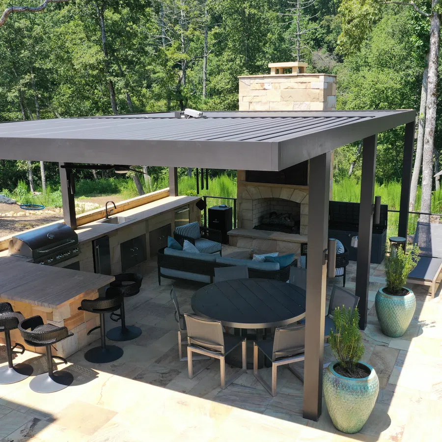 Modern outdoor patio with covered kitchen, bar stools, round dining table, fireplace, and lounge chairs surrounded by trees.