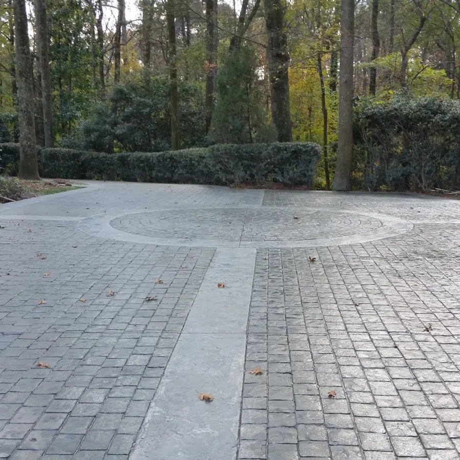 Stamped concrete driveway with decorative circular medallion and stone-pattern finish bordered by trees and shrubs in a wooded residential area.