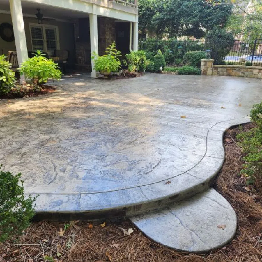 Curved stamped concrete patio with garden beds and plants under a covered porch in a backyard.
