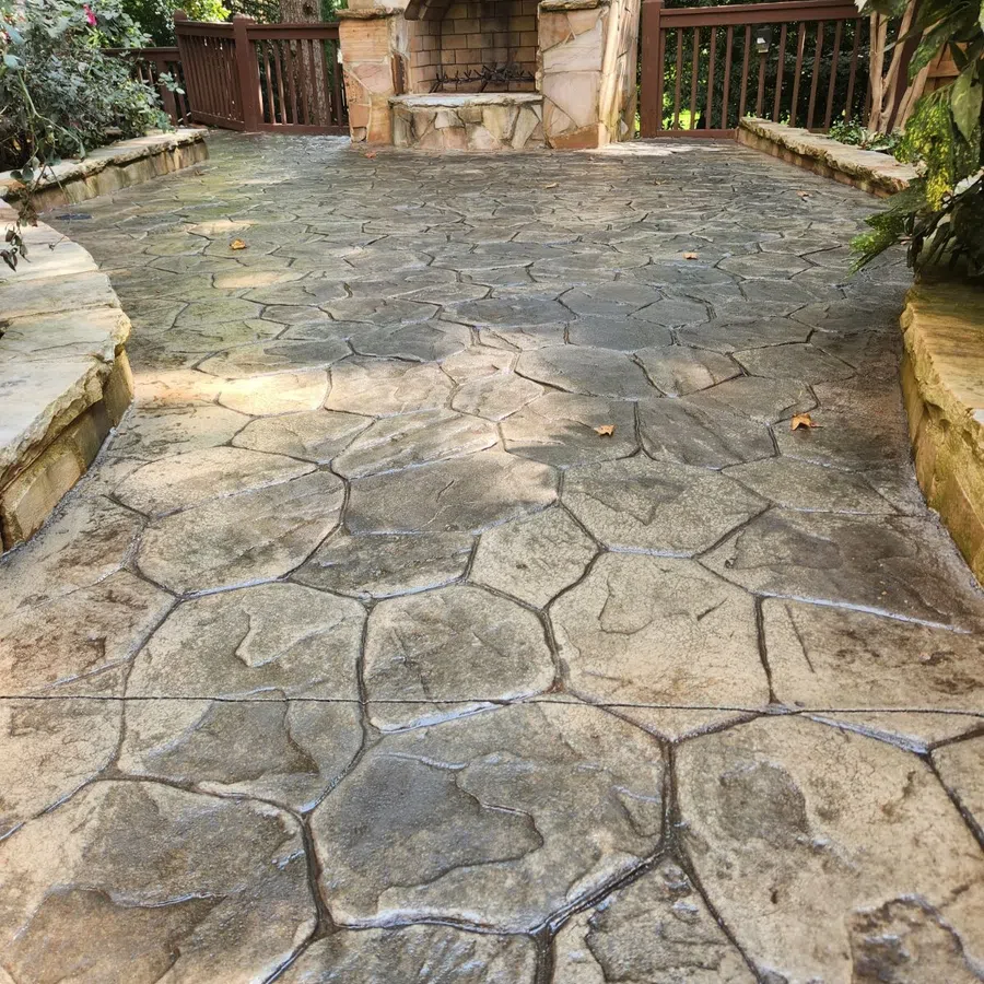 Outdoor stone patio with irregular patterned pavement and a stone fireplace surrounded by wood railing and greenery.