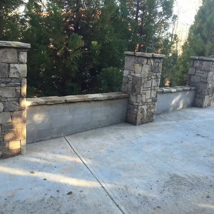 Stone pillars with low stone wall sections along a concrete driveway surrounded by greenery.