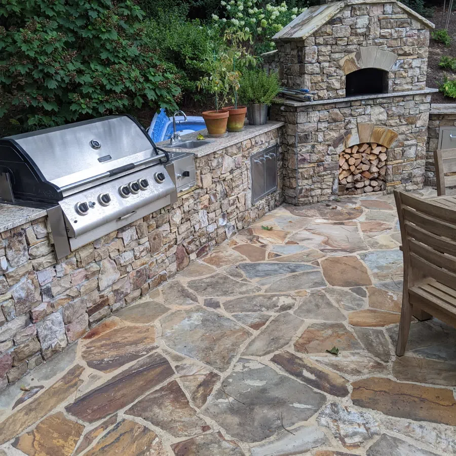 Outdoor stone kitchen with stainless steel grill, stone fireplace, potted plants, and natural stone patio flooring.