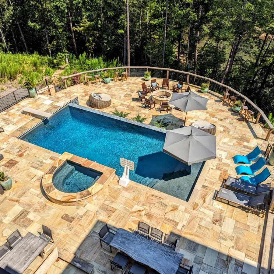 Aerial view of a backyard pool area with lounge chairs, umbrellas, fire pit, and natural stone patio surrounded by trees.