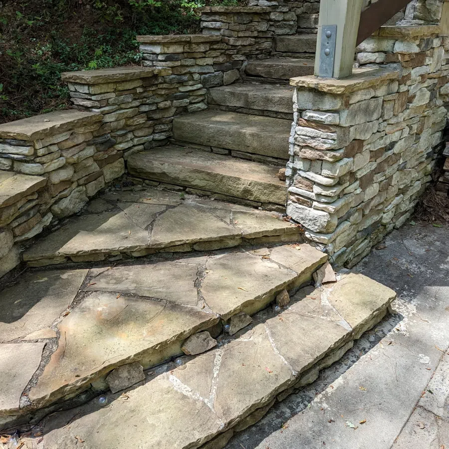 Curved stone staircase with natural stone walls and flagstone steps surrounded by greenery and concrete pathway.