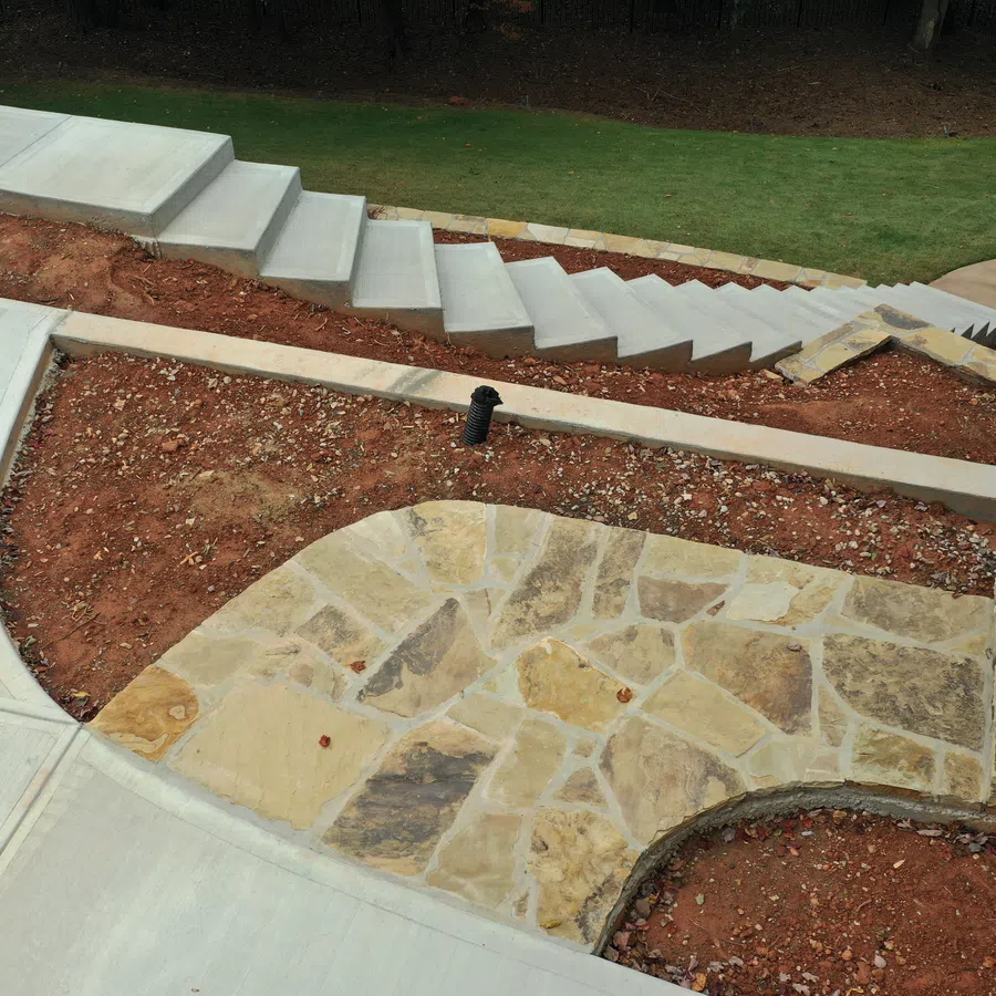 Outdoor stone patio with surrounding bare soil, concrete walkway, and stairway in a garden setting.