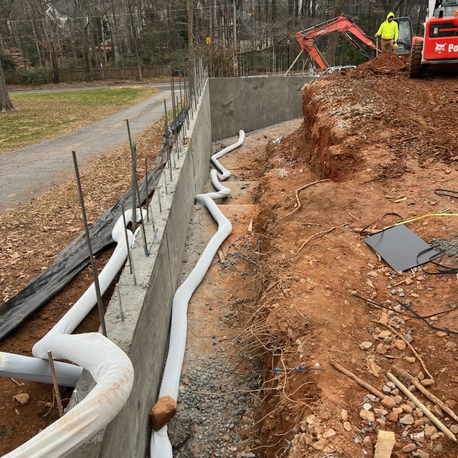 Construction site with concrete retaining wall, drainage pipes, exposed soil, and machinery working on earth excavation.