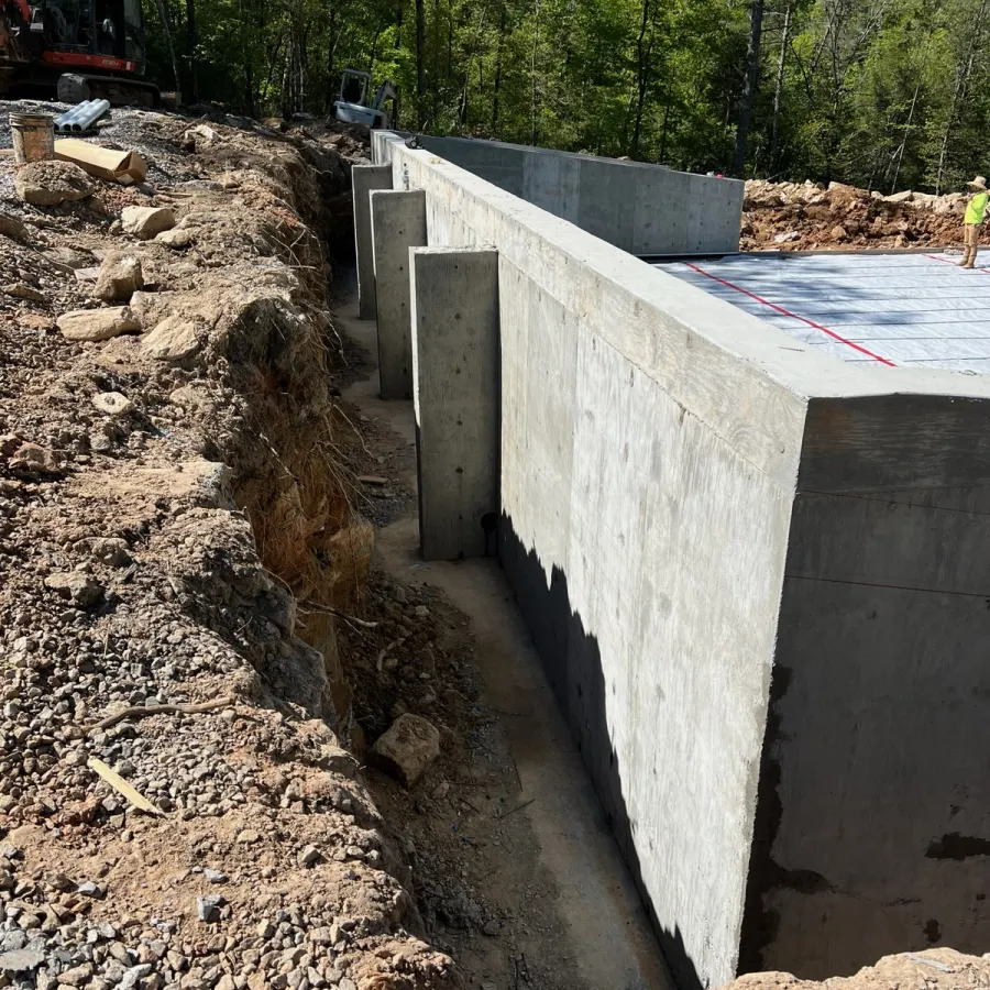 Concrete foundation walls with waterproofing layer in a construction site surrounded by trees and soil excavation.