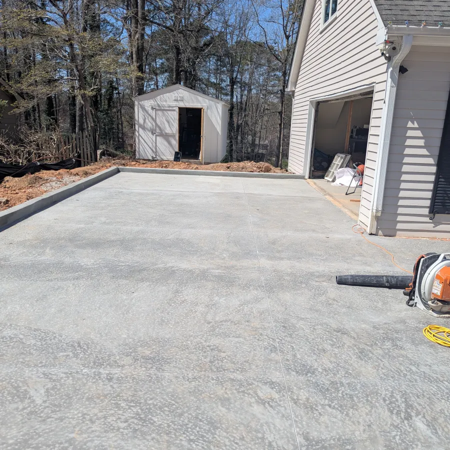 Freshly poured concrete driveway next to a white garage and a small white storage shed surrounded by trees.