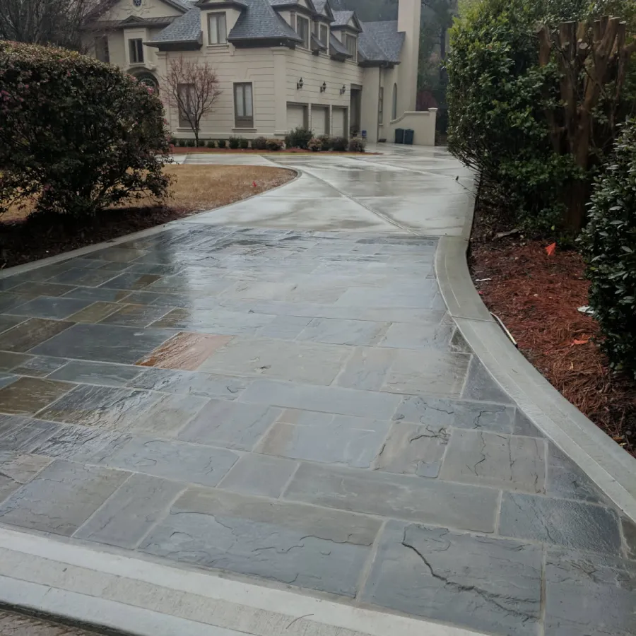 Wet stone driveway with concrete border leading to a large beige house surrounded by trees and shrubs.