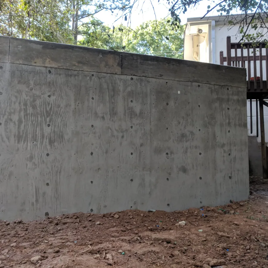 Newly poured concrete retaining wall in a backyard with dirt ground and wooden deck above