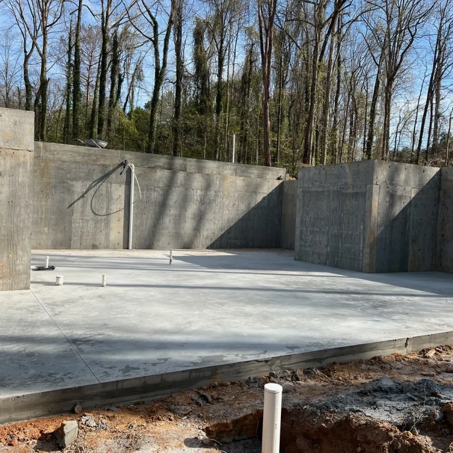 Concrete foundation and walls of a building under construction with surrounding trees and clear blue sky.