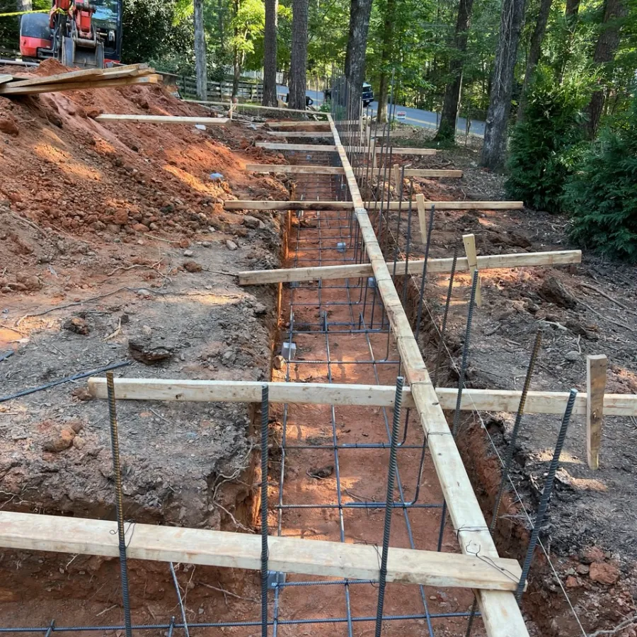 Construction site with wooden forms and rebar grid set in red dirt, surrounded by trees in a forested area.