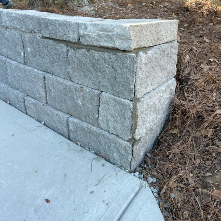 Close-up of a gray stone retaining wall next to a concrete sidewalk and dry ground with pine needles.