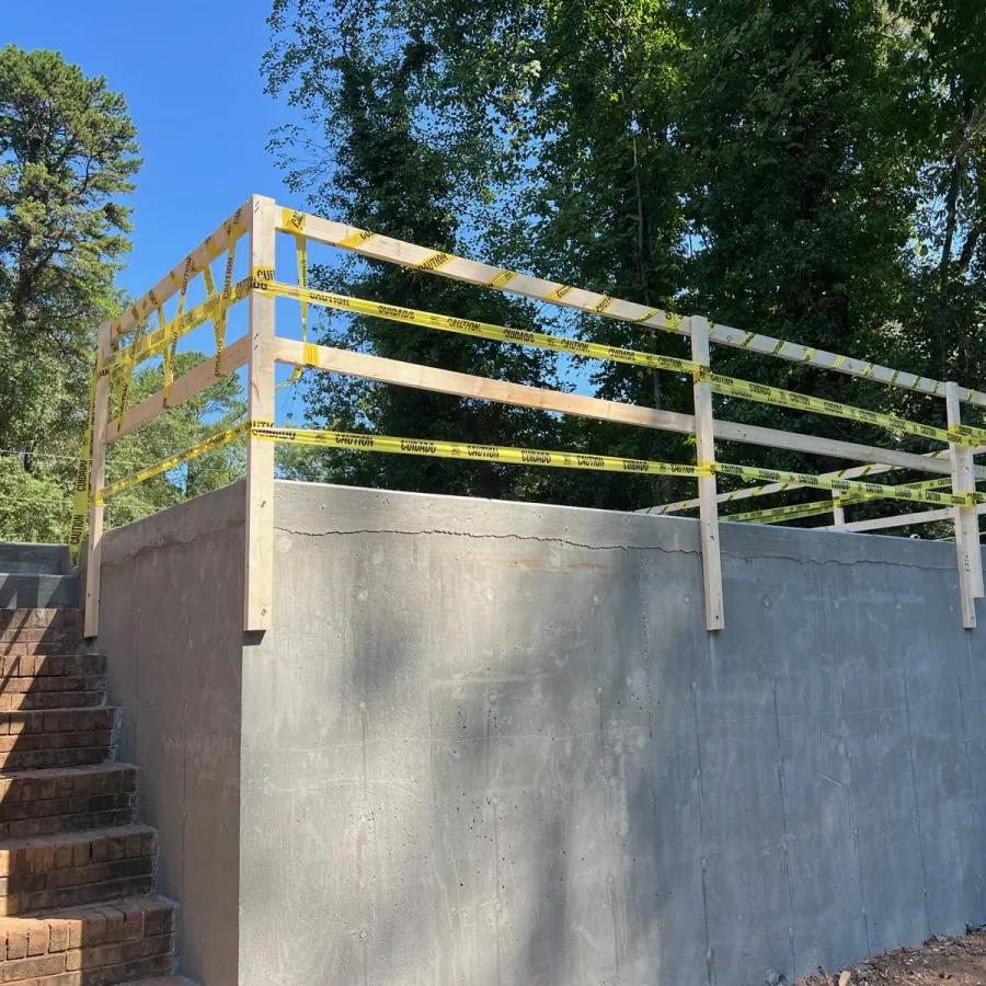Concrete platform with wooden railing and caution tape next to brick stairs surrounded by trees under blue sky.