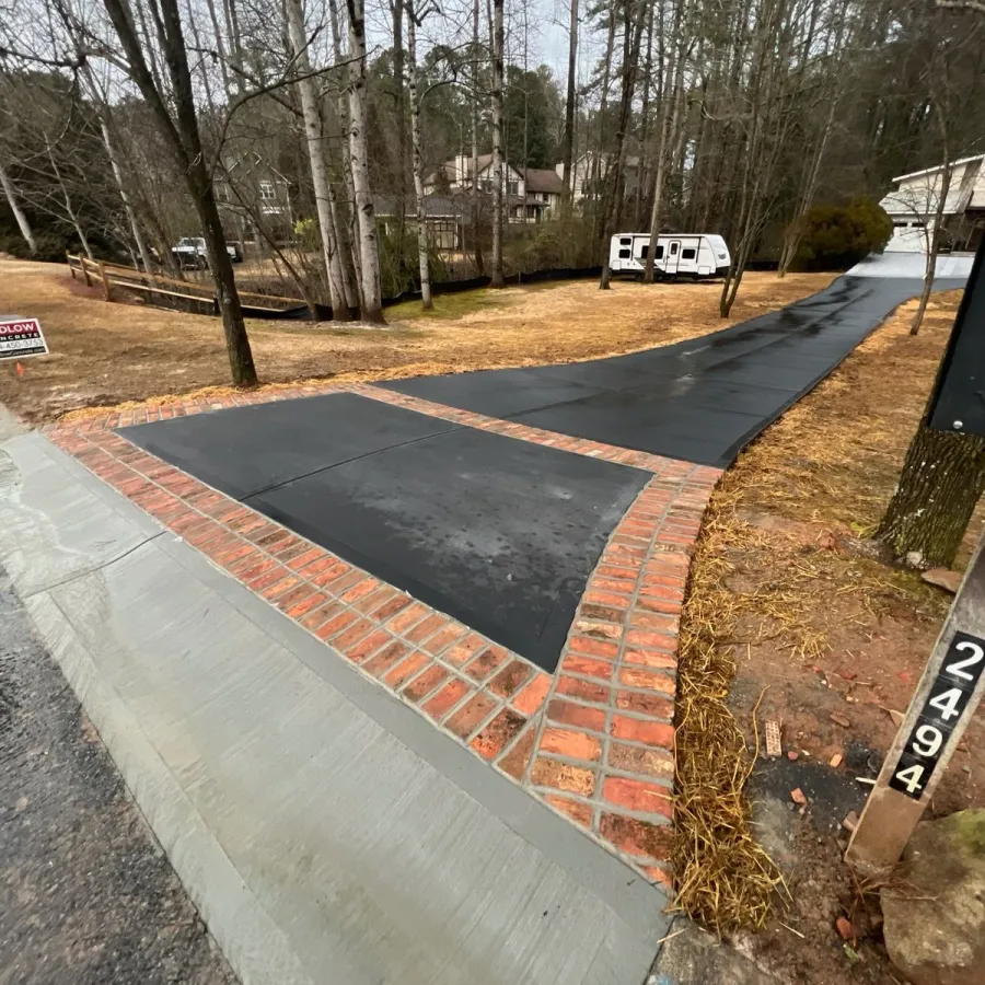 Newly paved black driveway with red brick border and concrete ramp leading to a house in a wooded area.