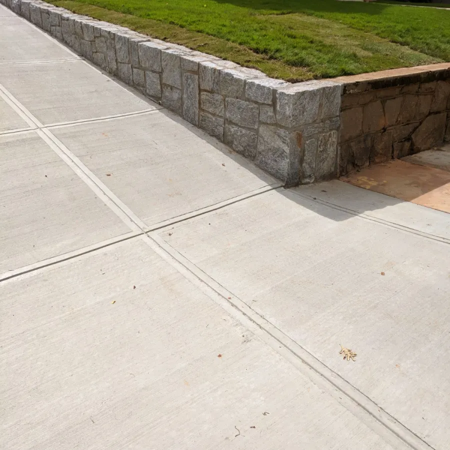 Clean concrete sidewalk next to a low stone retaining wall with green grass and house in background