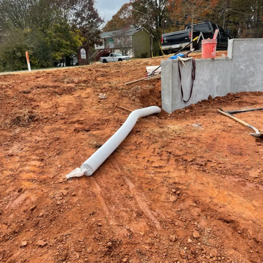 Construction site with exposed red soil, concrete wall, a white sediment control sock, and tools scattered around under cloudy sky.